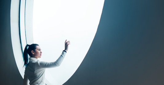 A woman stands gracefully in front of a large circular window, framed by natural light and soft shadows.